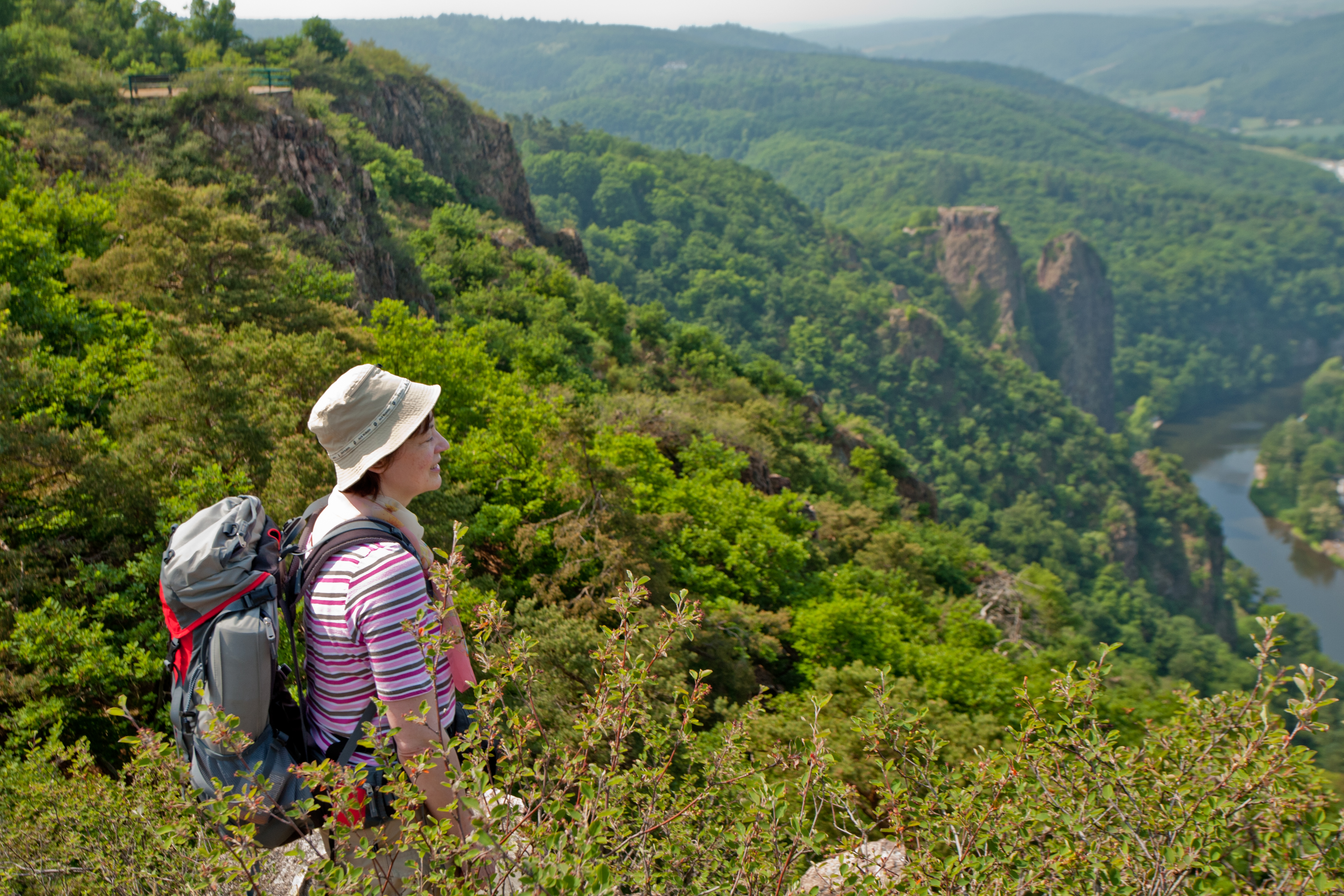 Frau mit Sonnenhut und Rucksack steht in bewaldetem Berghang und schaut in ein Flusstal und grünes Hügelland.